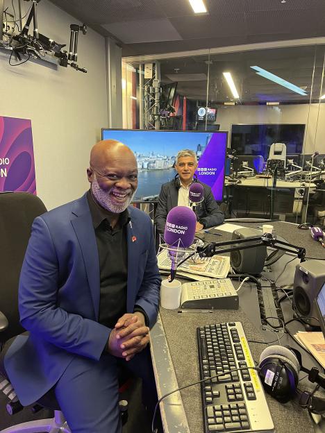 Eddie Nestor smiling in a radio studio with Sadiq Khan in the background.