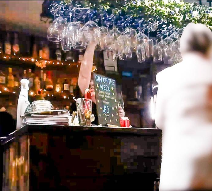 A person standing at a bar with glasses hanging overhead and a "Gin of the Week" sign visible.