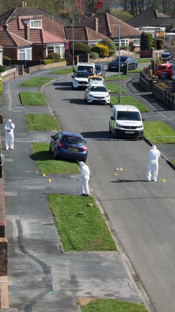 A crime scene with forensic investigators in white suits and police tape around a blue car in a residential street.