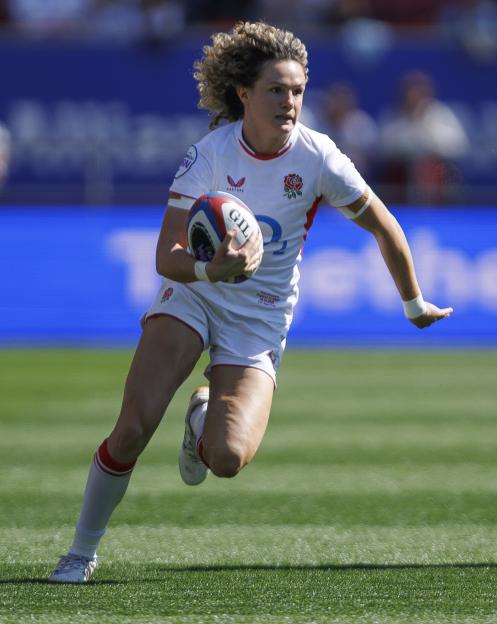 A female rugby player in an England jersey runs with the ball on a green field.