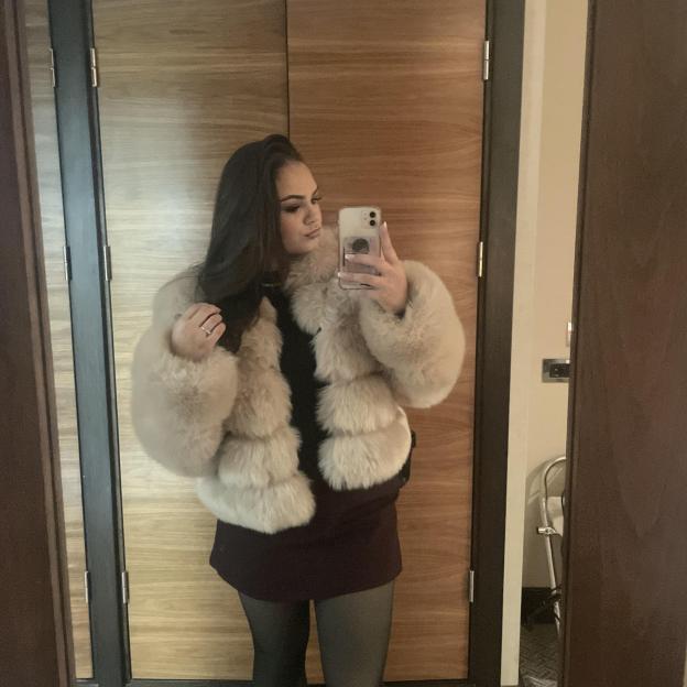 A woman in a fur coat, dark dress, and tights takes a mirror selfie.