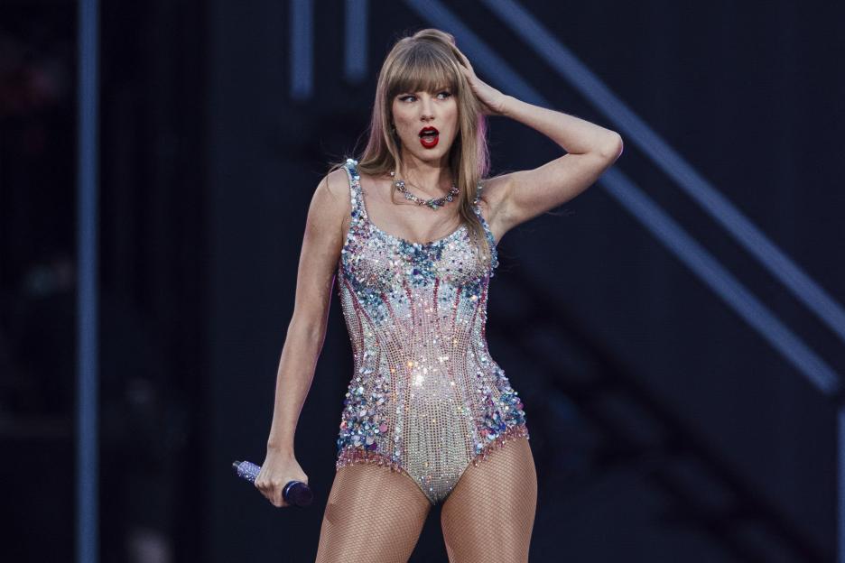 Taylor Swift performing on stage in a sequined costume and fishnet stockings, holding a microphone, with her hand on her head and mouth open as if singing.