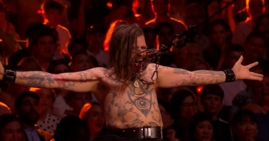 A male performer with dreadlocks and tattoos on his chest and arms is chained with his arms outstretched while a large chain is wrapped around his face, with an audience in the background.