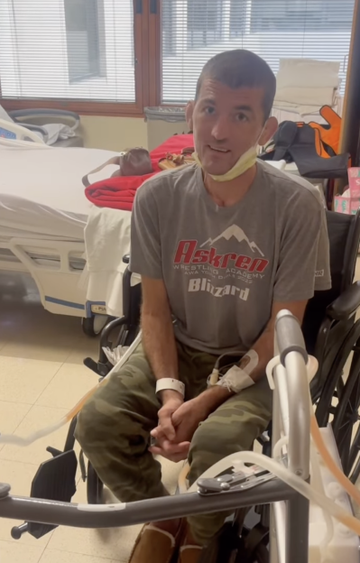 A man in a hospital, wearing an Askren Wrestling Academy Blizzard t-shirt, sits in a wheelchair with IVs in his arms.
