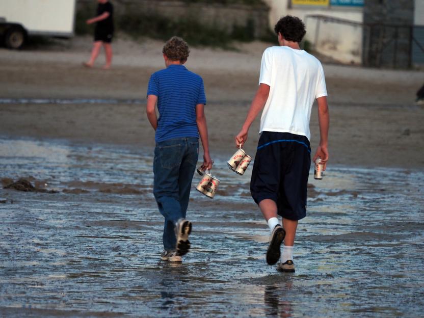 Two young men carry several beer cans as they walk across a wet, sandy beach.
