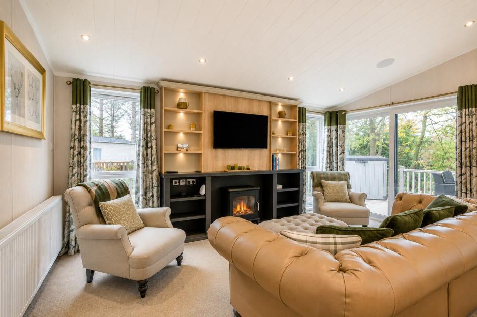 Living room with a large tan leather couch, two armchairs, a television mounted above a fireplace, and large windows looking out to trees.