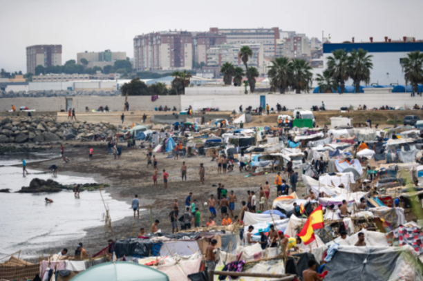A crowded beach in Ceuta, Spain, with makeshift tents and people by the water, with city buildings in the background.