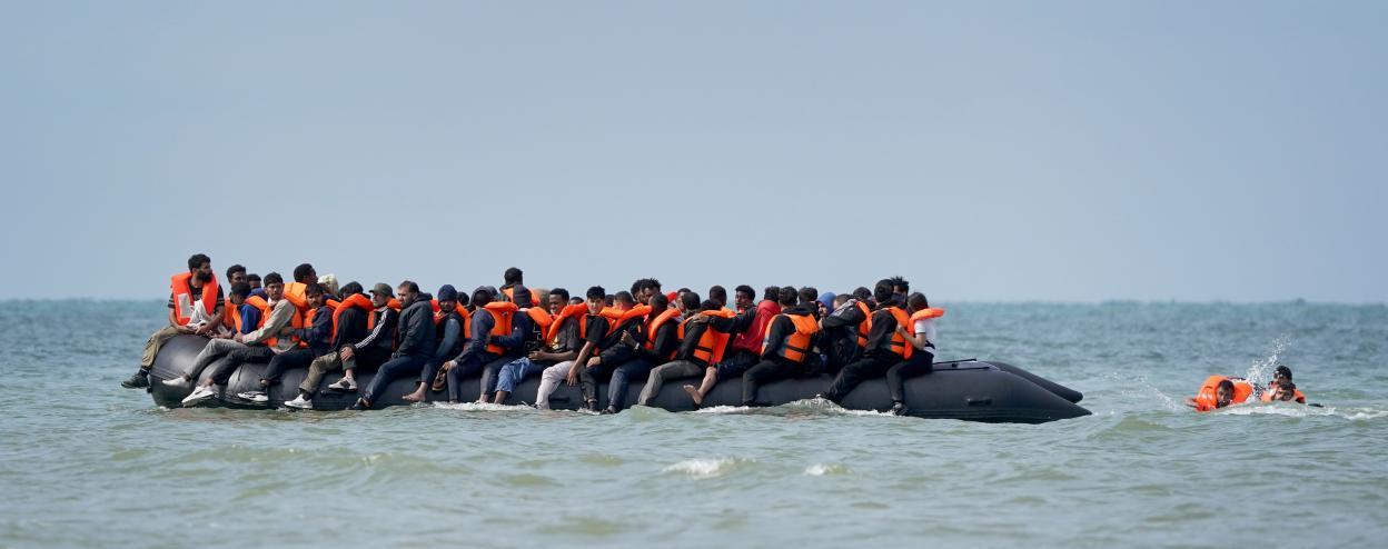 People in orange life vests board a small, black inflatable boat on the sea near Equihen-Plage, France, with others swimming nearby.