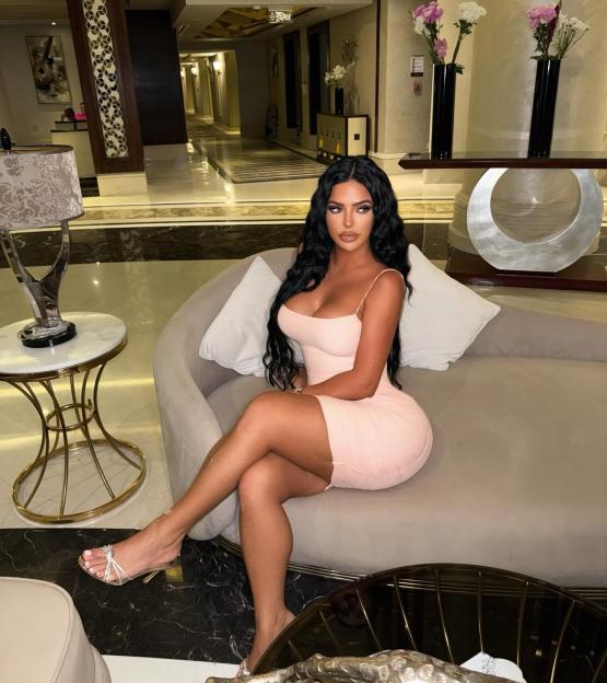 A woman with long dark hair and a light pink dress sits on a gray sofa in a hotel lobby.