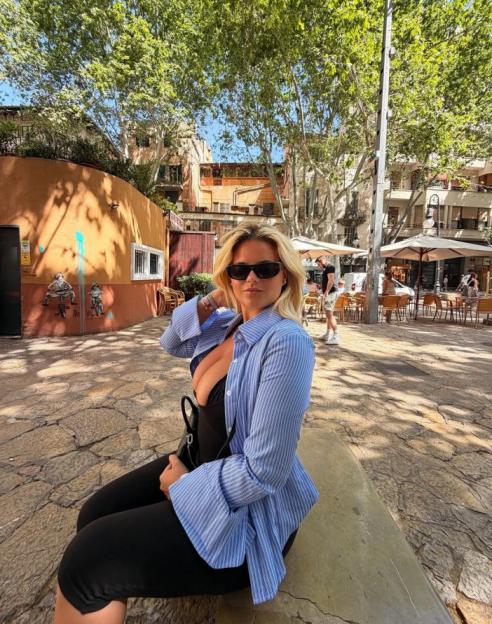 Woman in a black dress, blue striped shirt, and sunglasses sitting on a stone bench outdoors.