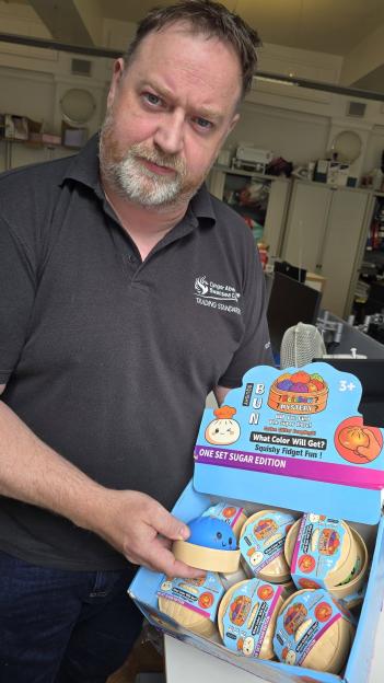 A man in a black polo shirt holding a "squishy dumpling" toy from a box containing several similar toys.