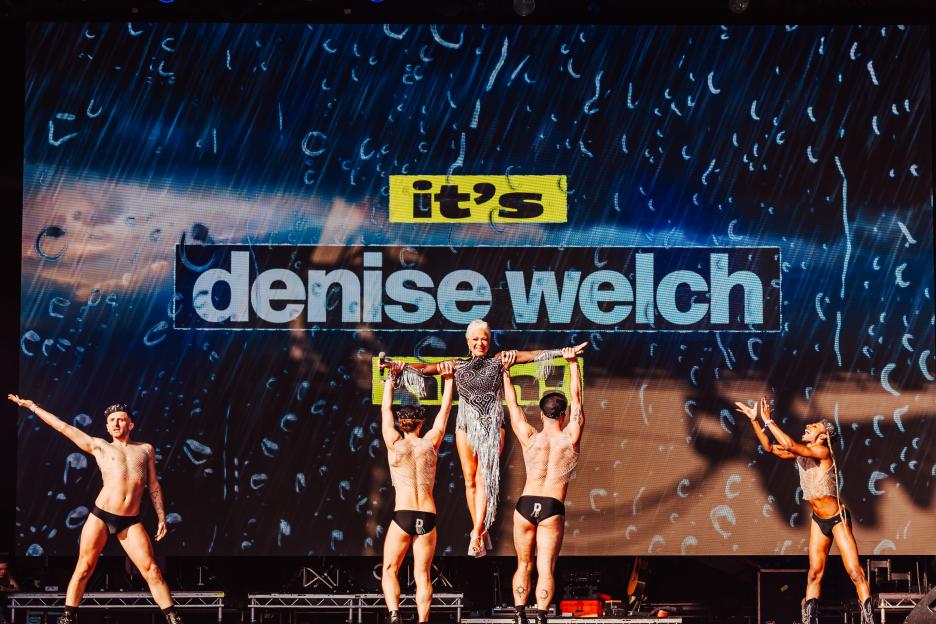 Denise Welch performing at Mighty Hoopla, held up by two dancers, with a large screen behind her displaying "it's denise welch" and a rain effect.