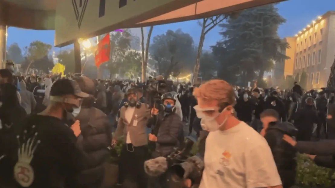 A large crowd gathers outdoors at dusk, with several individuals in the foreground wearing masks and protective gear.