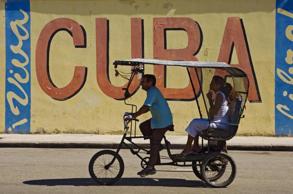 Two people ride in a pedicab with a "Viva Cuba" mural in the background.