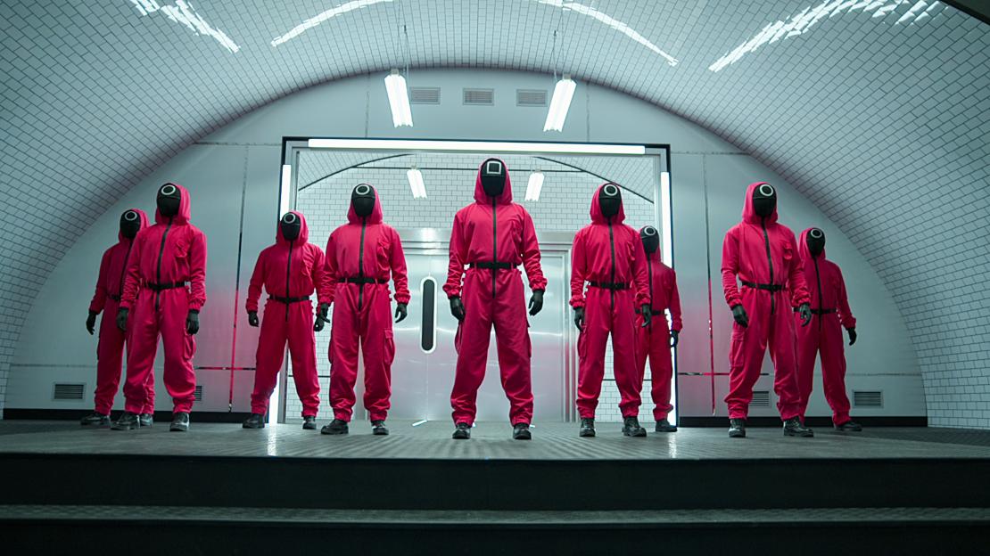 Eight guards in red jumpsuits and black masks stand on a raised platform under white subway tiles.