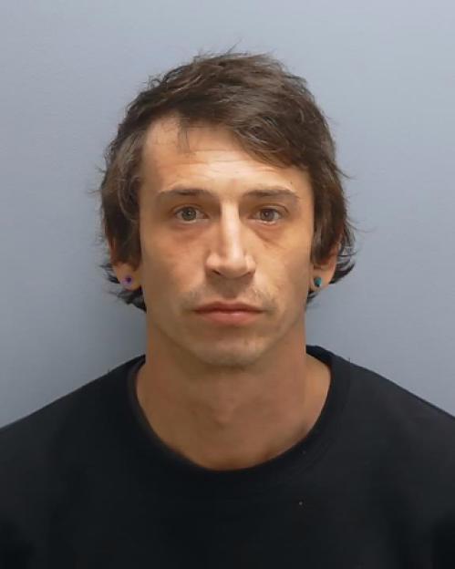 Mugshot of a man with dark hair, wearing a black shirt, looking directly at the camera.