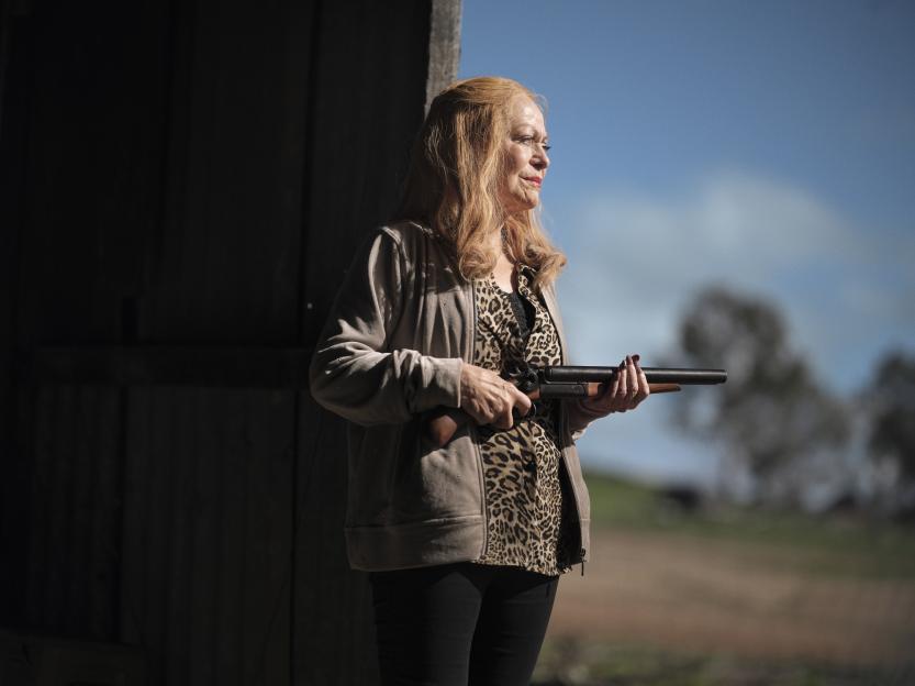 A woman with red hair, wearing a leopard print top and a light brown jacket, holding a shotgun and looking into the distance.