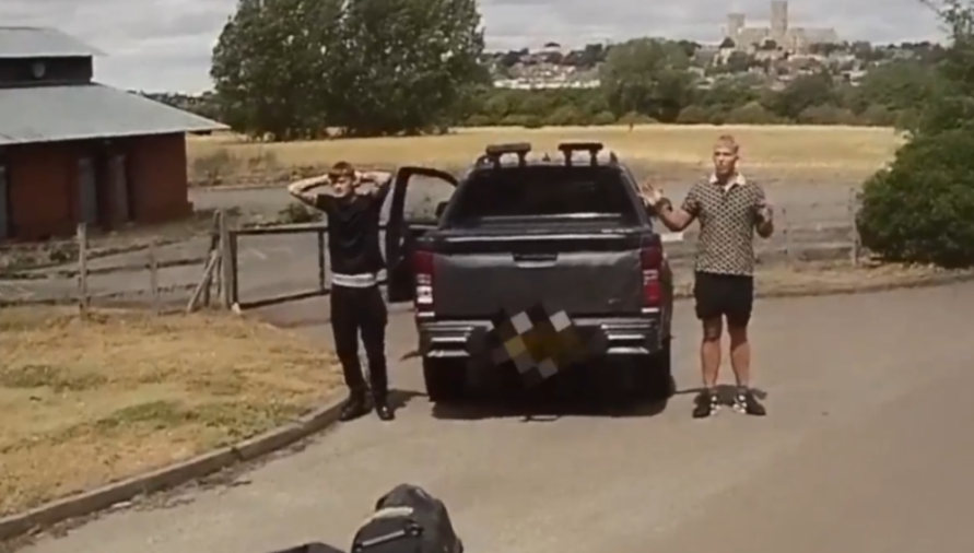 Two men with their hands up standing near a dark pickup truck, while a medieval castle is visible in the background.