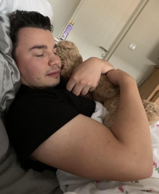 A man with dark hair sleeps in bed, cuddling a teddy bear.