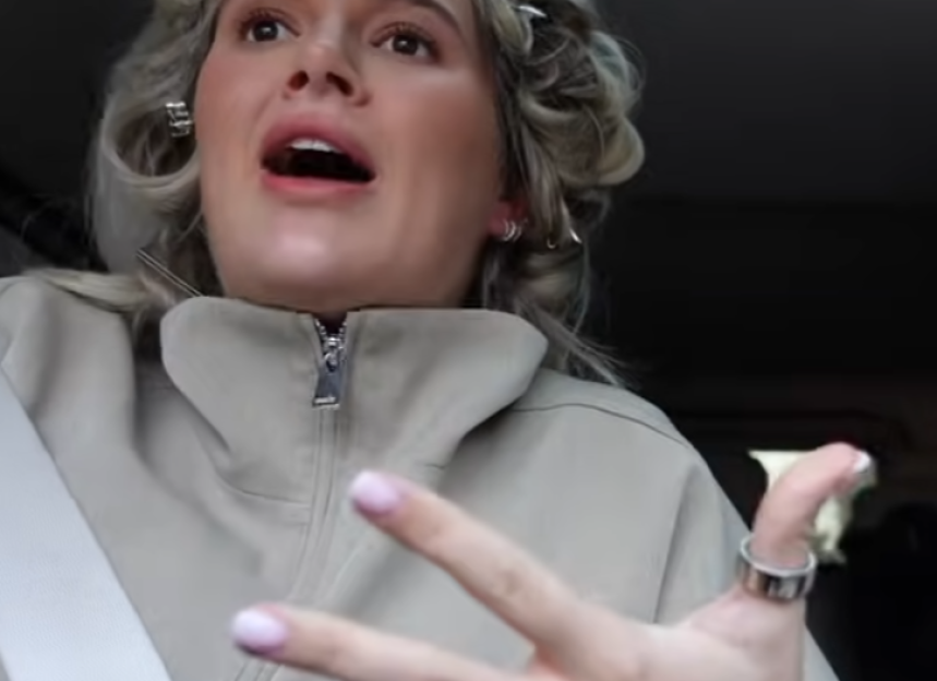 A woman in a beige jacket with her hair in rollers talks with her mouth open and gestures with her right hand.