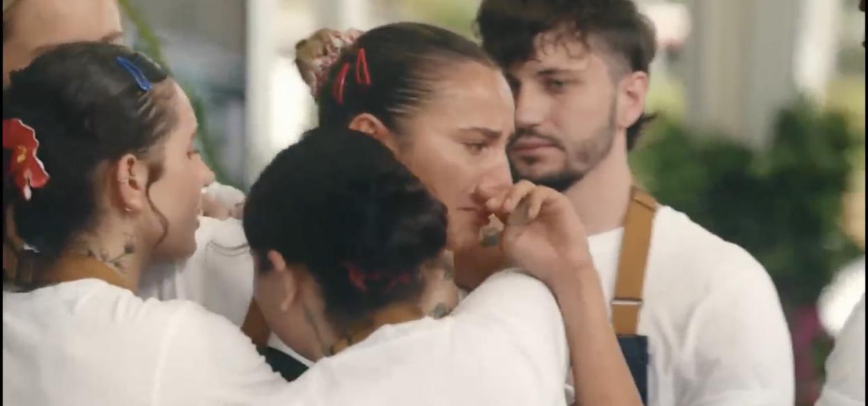 A woman in a white t-shirt and red hair clips crying and being consoled by others.