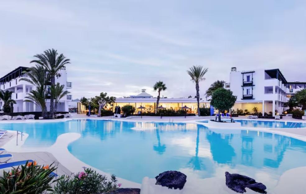 A holiday resort with a large swimming pool, palm trees, and buildings with balconies.