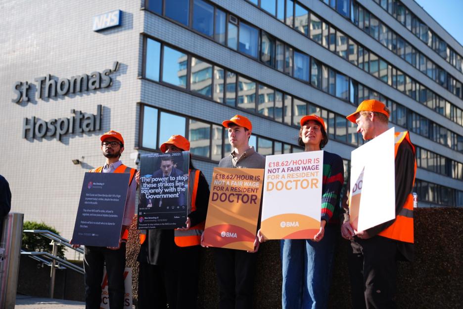 Resident doctors on strike outside St. Thomas' Hospital holding signs that indicate £18.62/hour is not a fair wage for a resident doctor.
