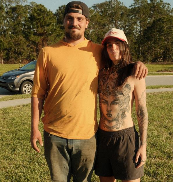 A person with long hair and elaborate chest tattoos stands next to a man with his arm around them.