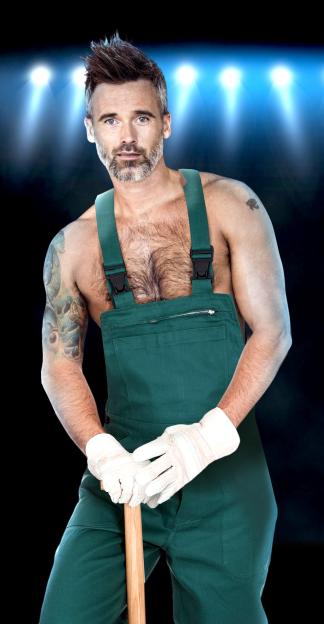 A man with spiky grey hair, a beard, and a hairy chest wears green overalls and work gloves, holding a wooden handle, against a dark background with blue spotlights.