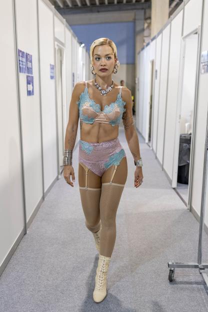 Rita Ora walking in a hallway, dressed in a pastel pink and blue lace bra and underwear, with fishnet stockings and cream-colored lace-up boots.