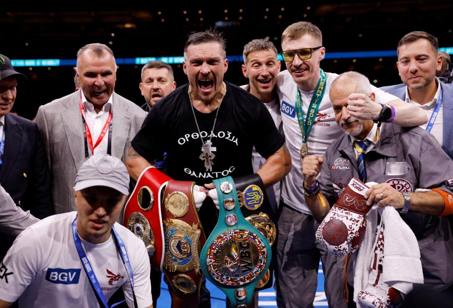 Oleksandr Usyk celebrates winning his fight against Daniel Dubois, surrounded by his team, holding multiple championship belts.