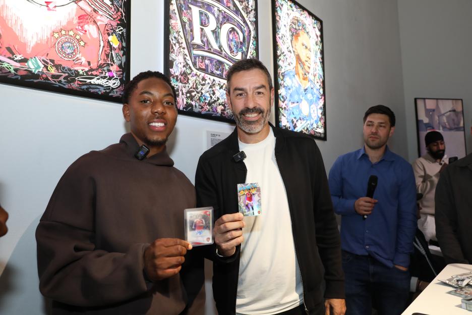 Two men holding sports trading cards pose in front of abstract art.