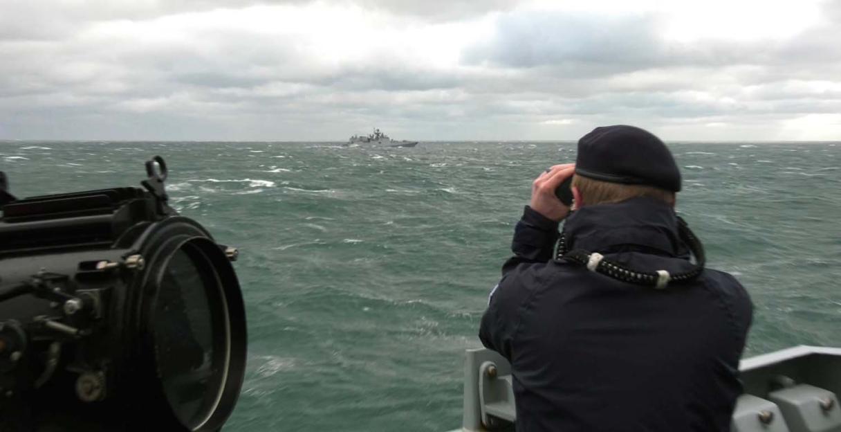 A Royal Navy officer monitors a Russian warship with binoculars on a stormy sea.