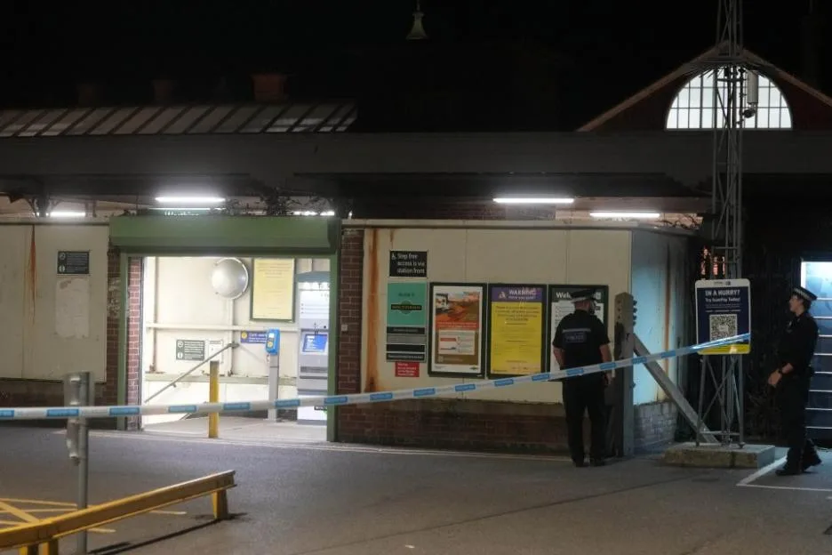 Police officers stand guard behind police tape outside a closed Worthing station at night.