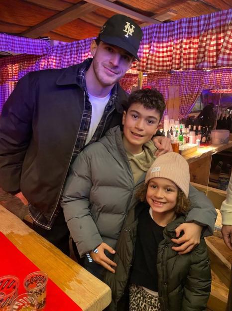 A man in a black jacket and baseball cap with his arms around two children, one boy and one girl, all smiling at the camera.