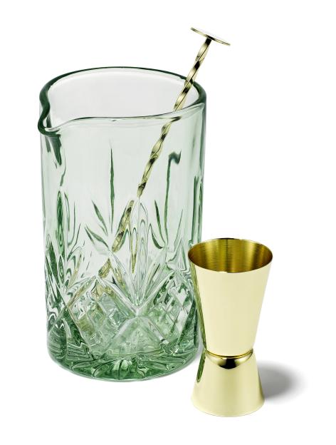 A green glass mixing jug with a gold bar spoon and a gold jigger.