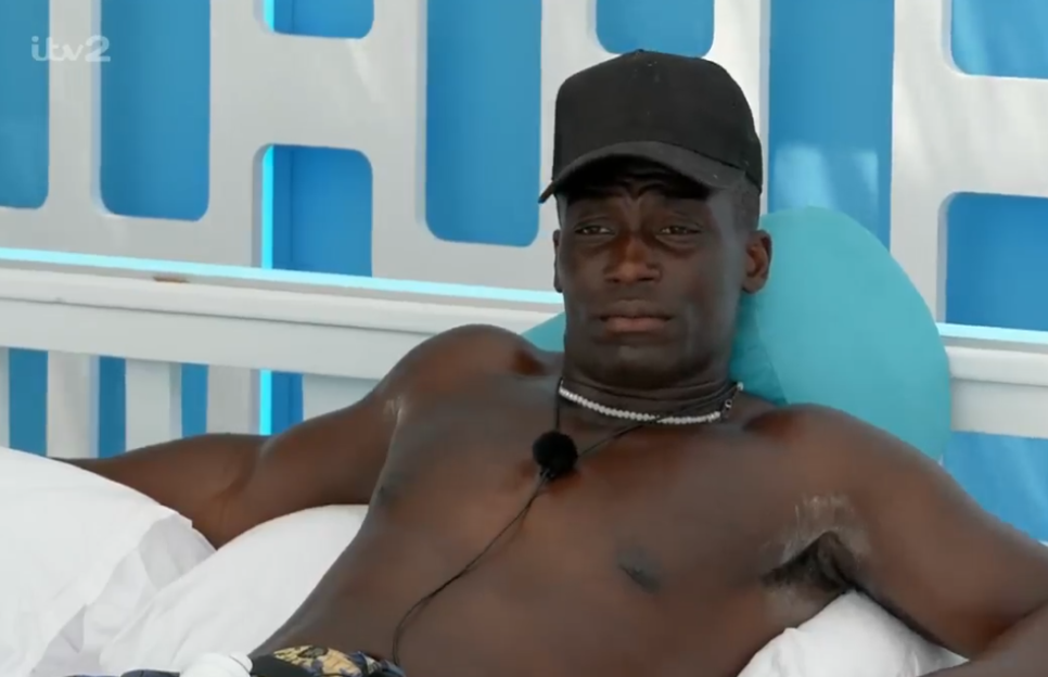 A man in a black cap and necklace sits shirtless, looking at the camera.