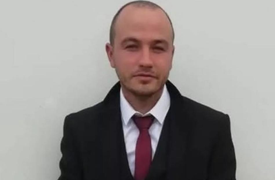Headshot of a man with a short beard and bald head wearing a black coat, white shirt, and red tie.
