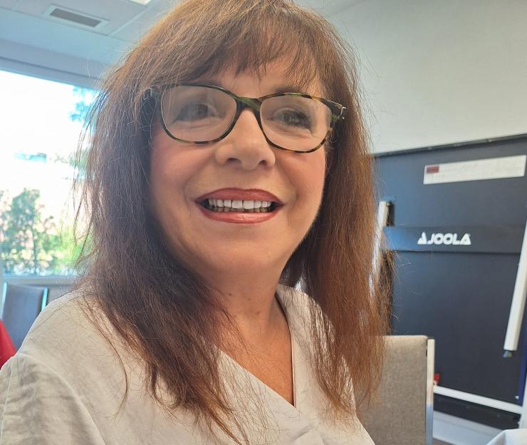 A smiling woman with reddish-brown hair and glasses wearing a white shirt and blue pants.