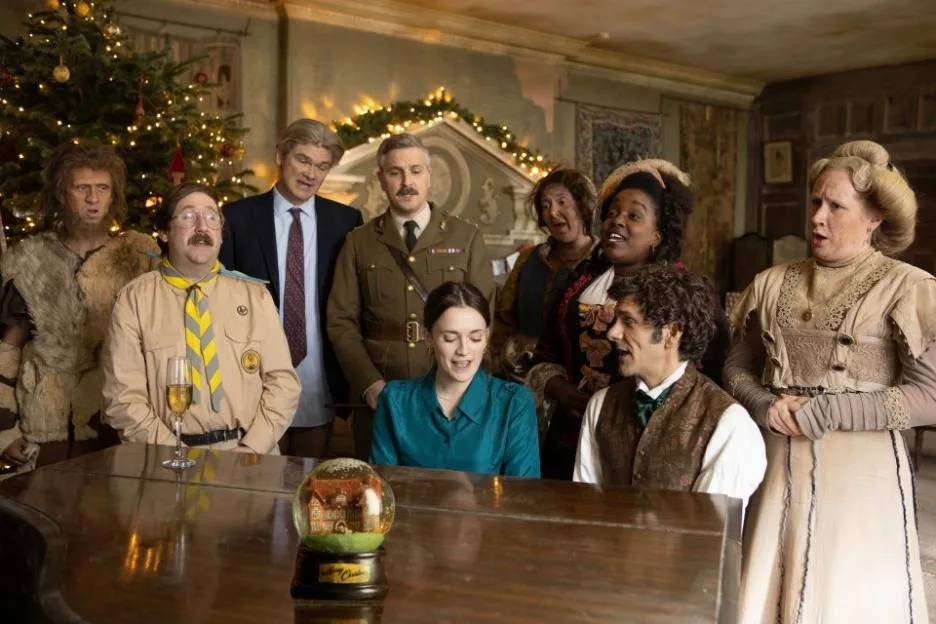 The cast of the TV show Ghosts gather around a piano to sing in a festively decorated room.