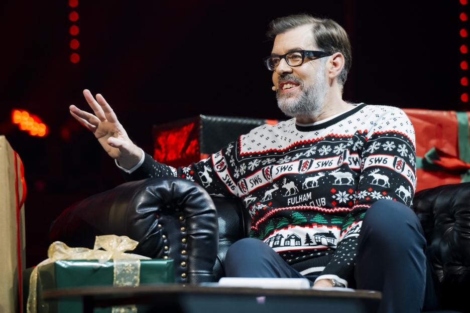 Richard Osman wearing a festive "Fulham Club" sweater and glasses, smiling while sitting on a black leather couch, gesturing with his left hand.