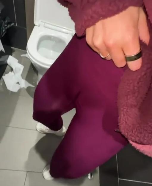 A person in purple leggings and a maroon jacket stands in a bathroom with a white toilet in the background.