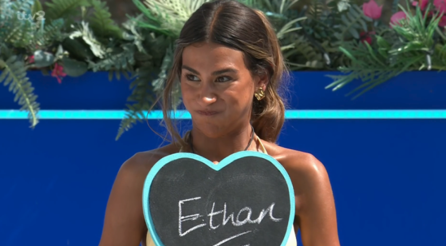 A woman in a cream top holding a heart-shaped chalkboard with "Ethan" written on it.