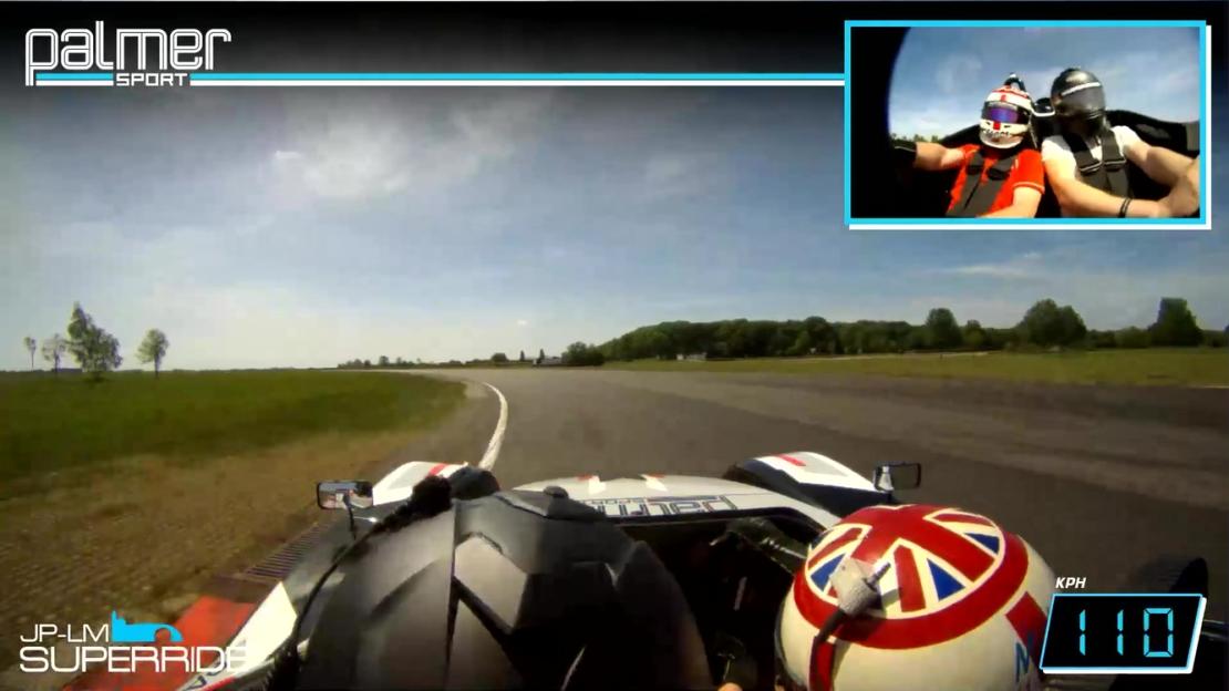 POV shot from a race car on a track, with a small inset showing two people in a car.