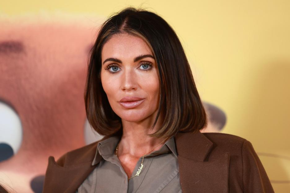 A woman with shoulder-length dark hair and blue eyes, wearing a brown coat and gray collared shirt, and a gold necklace with the name "Ritchie."