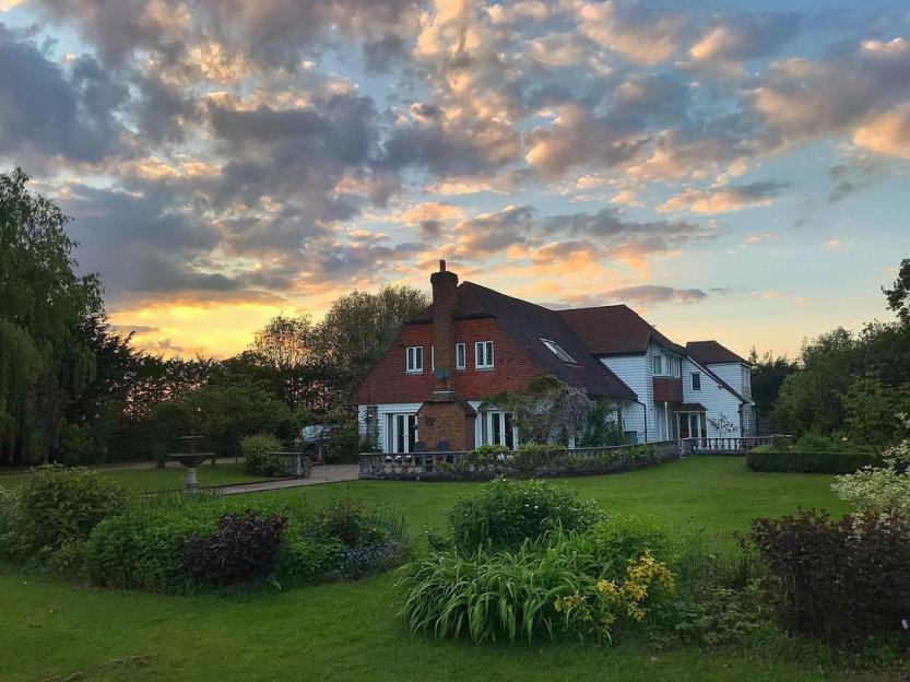 A large house with a red tiled roof and white siding, surrounded by a green lawn and gardens under a sunset sky.