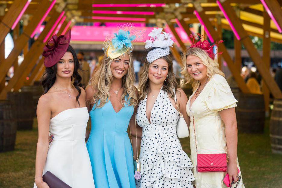 Four women in dresses and fascinator hats smiling.