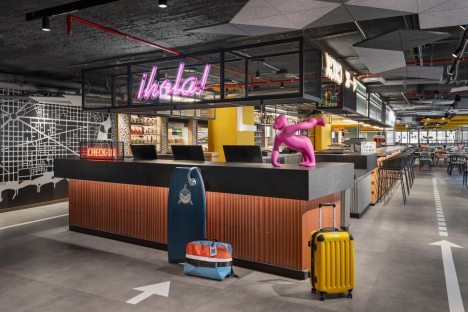 The check-in desk at Moxy Barcelona Hotel with a pink gorilla statue, "¡hola!" neon sign, and "CHECK-IN" sign, and a map of Barcelona on the wall.