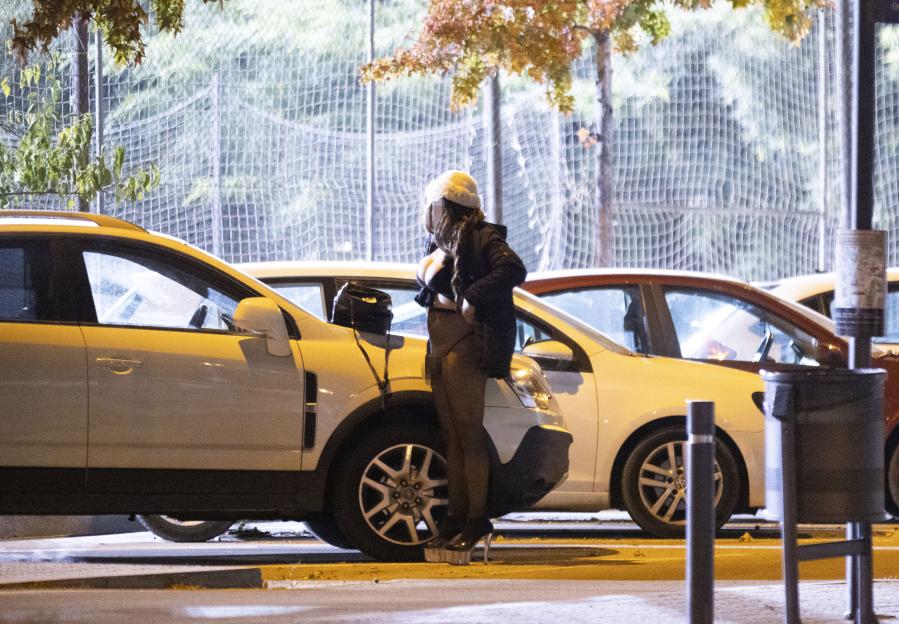 A woman in a white hat, black top, and dark leggings stands in high heels beside a parked white car at night.