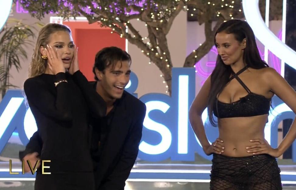 A woman with blonde hair covers her face with her hands, looking shocked, while a man in a suit smiles and hugs her from behind, and another woman in a black crop top stands nearby with hands on hips and a slight smile.